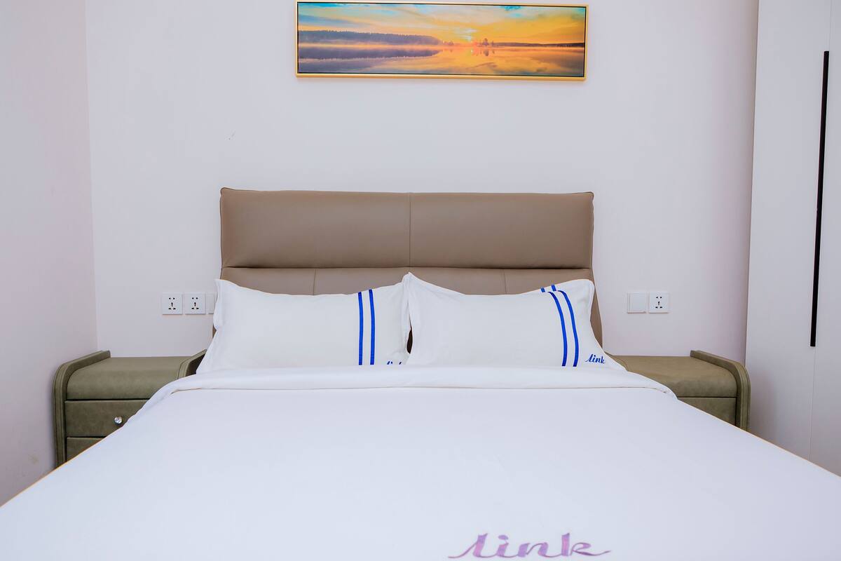 A neatly made bed with white bedding and blue stripes, a brown headboard, and a painting of a sunset on the wall.