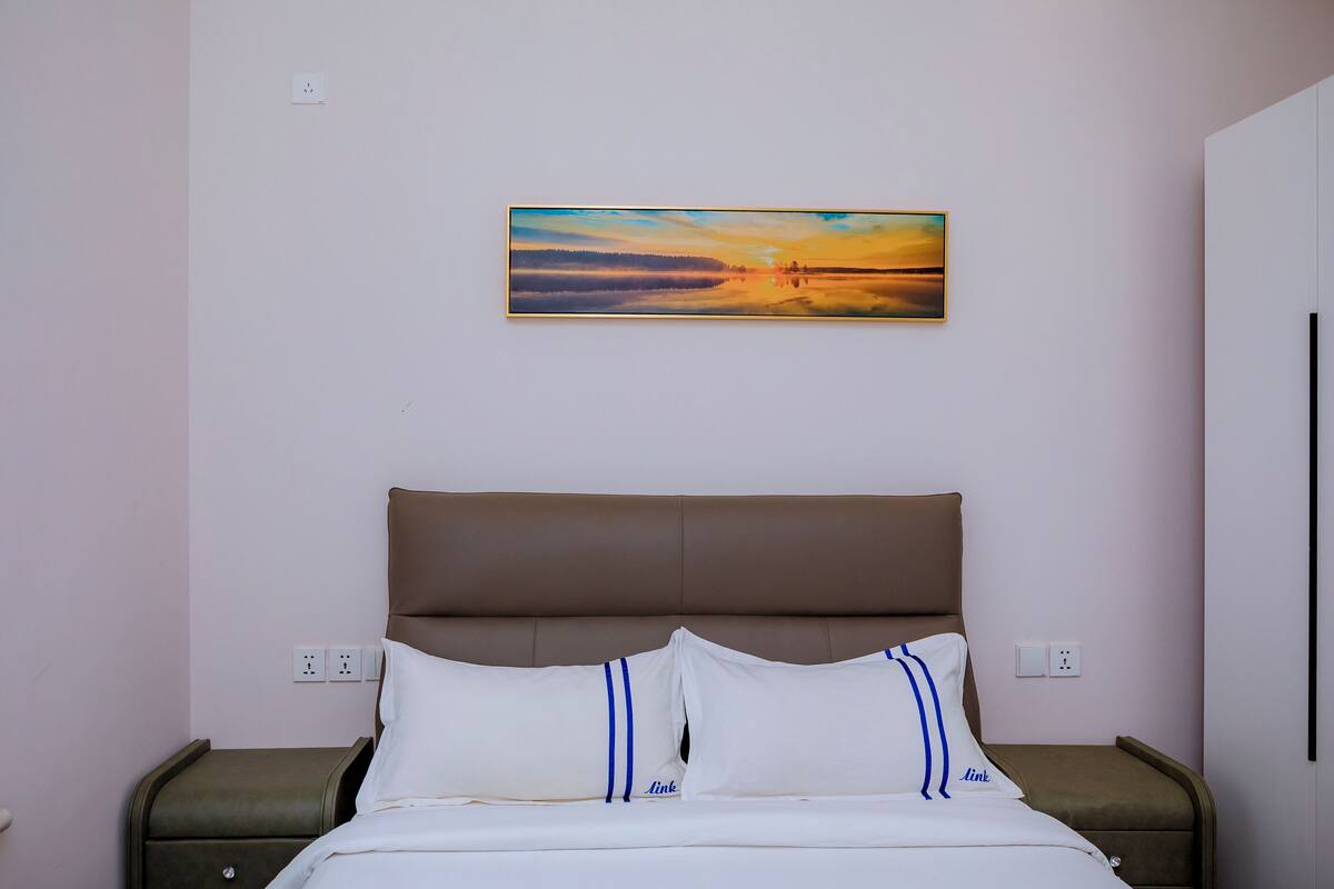 A bedroom with a bed, a headboard, and a painting of a sunset on the wall.