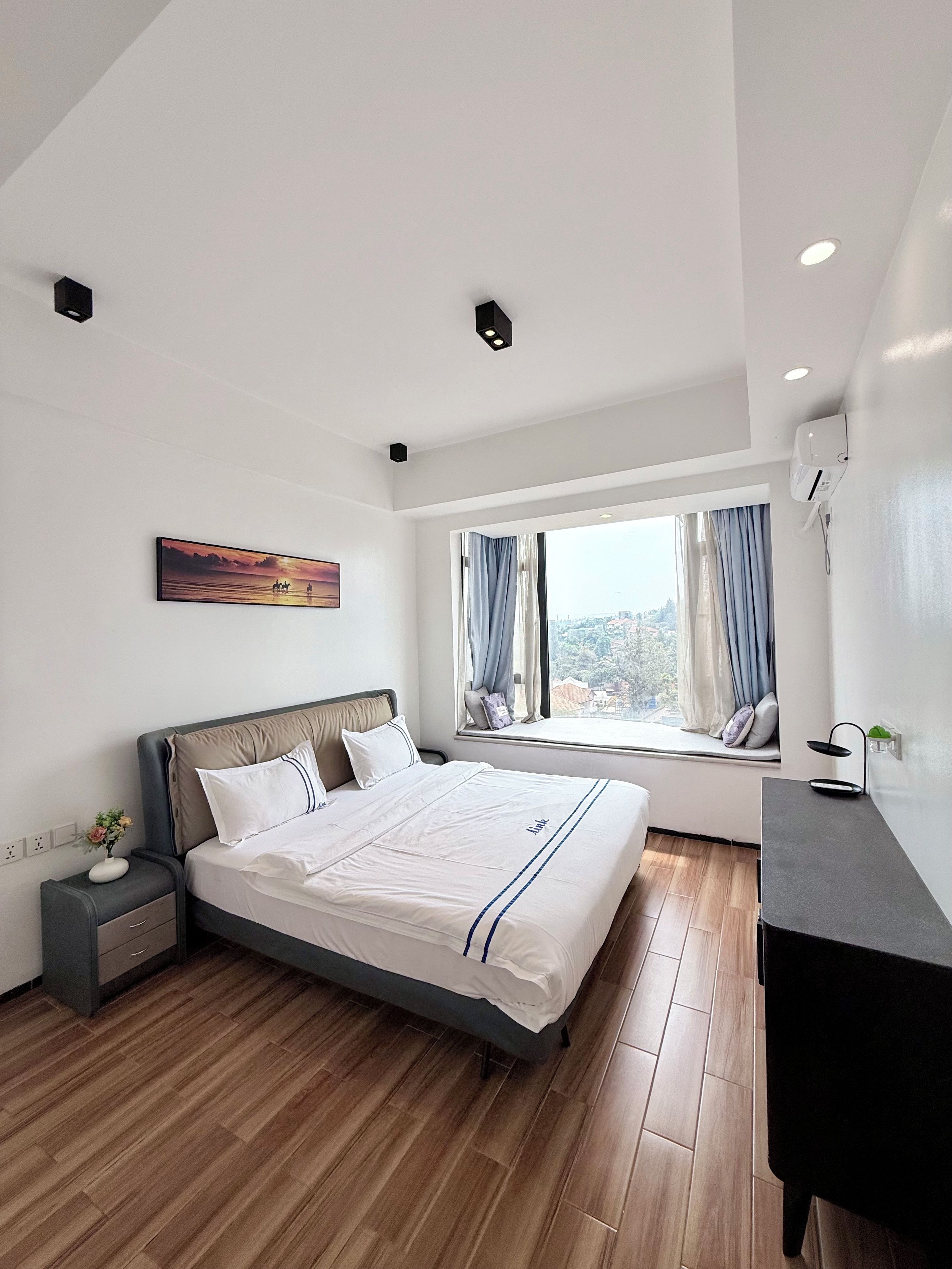 A modern bedroom with a large bed, a bedside table, a nightstand, a window with curtains, and a painting on the wall.