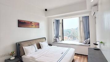 A modern bedroom with a large bed, a bedside table, a nightstand, a window with curtains, and a painting on the wall.