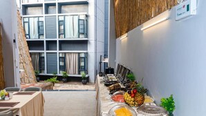 Disposición de un bufé de desayuno en un hotel con pared de piedra, vista a un edificio de varios pisos y diversas opciones de comida dispuestas sobre una mesa.