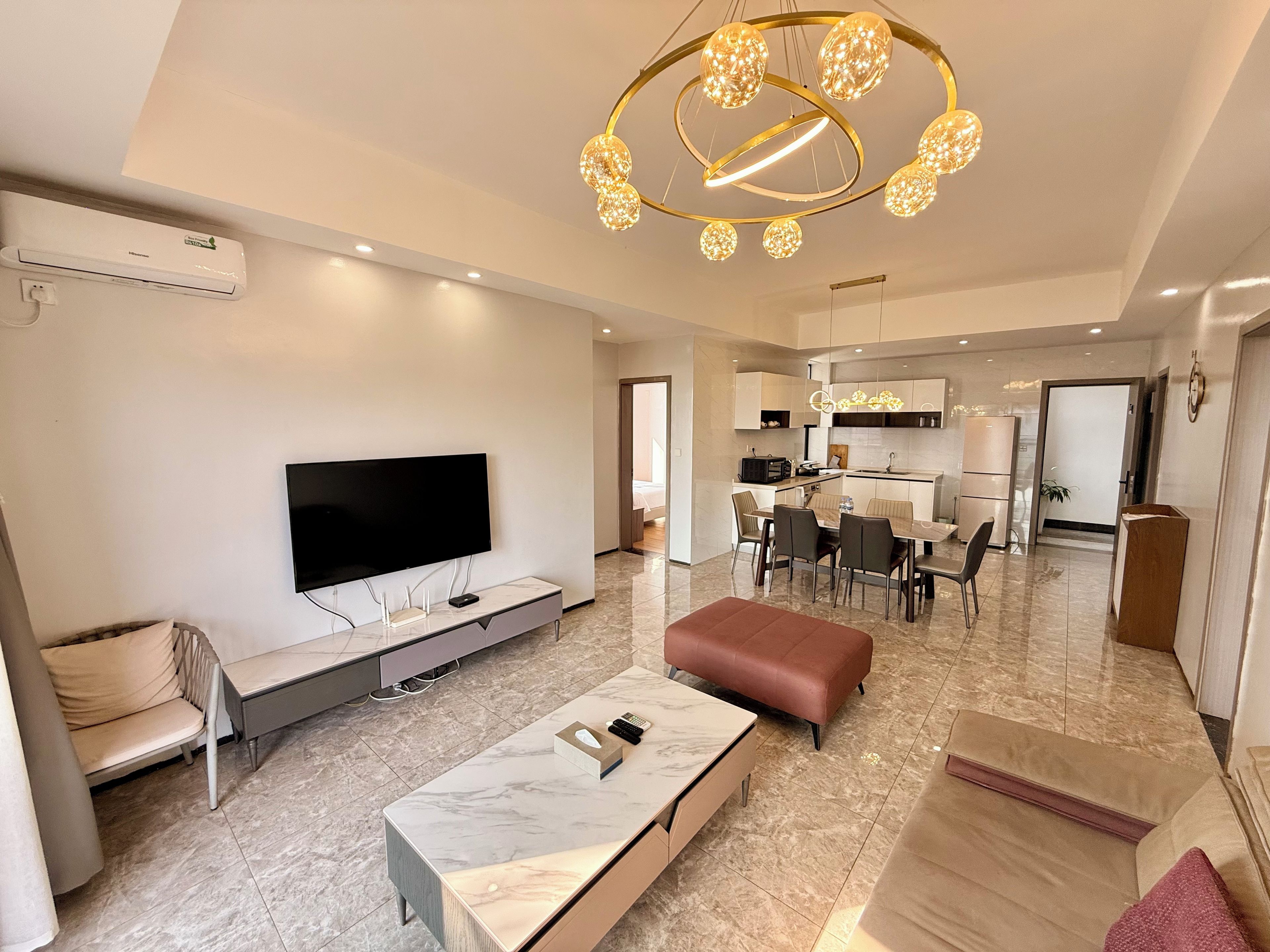 A modern living room with a flat-screen TV, a marble coffee table, and a sofa. There is a dining area with a table and chairs, and a kitchen in the background.