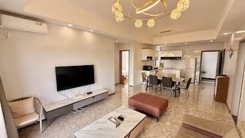 Una sala moderna con un televisor de pantalla plana, una mesa de centro de mármol y un sofá. Se observa un comedor con una mesa y sillas, y una cocina al fondo.