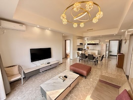 Una sala moderna con un televisor de pantalla plana, una mesa de centro de mármol y un sofá. Se observa un comedor con una mesa y sillas, y una cocina al fondo.