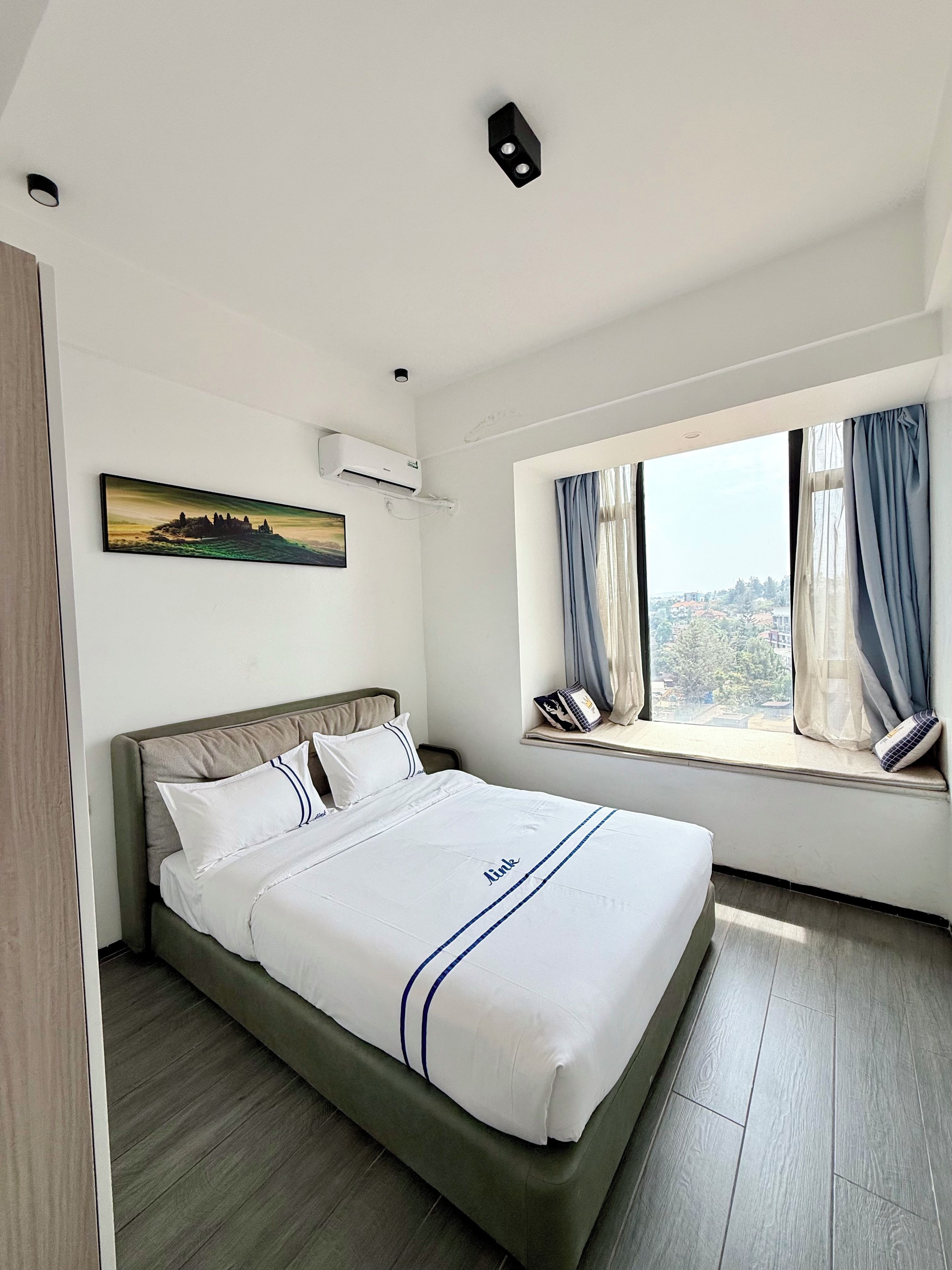 A hotel room with a bed, a window with curtains, an air conditioning unit, and a painting on the wall.