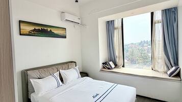 A hotel room with a bed, a window with curtains, an air conditioning unit, and a painting on the wall.