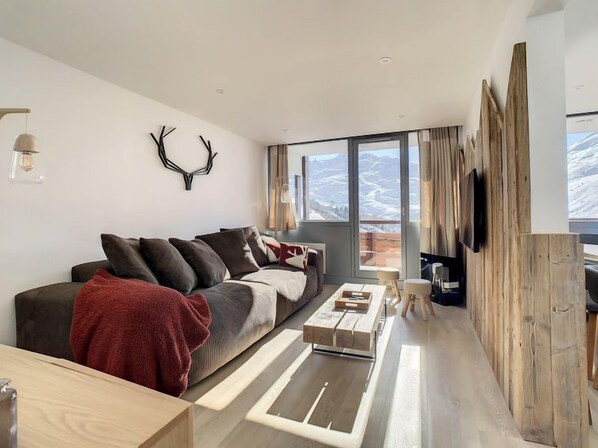A modern living room with a large window, a sofa, a coffee table, and a TV mounted on a wooden wall.