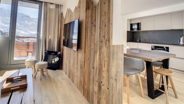 Eine moderne Küche mit Holzelementen, ein Essbereich mit Holztisch und -stühlen sowie ein großes Fenster mit Blick auf schneebedeckte Berge.