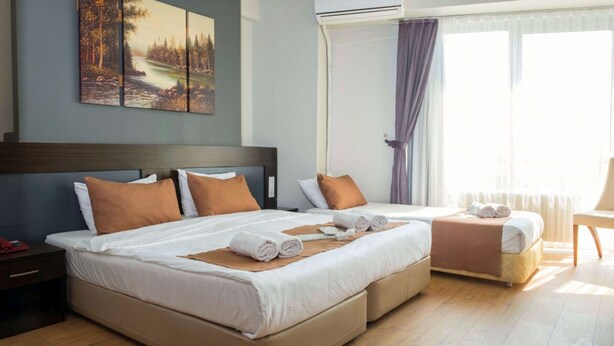A hotel room with two beds, a large window with curtains, a painting on the wall, and a bedside table with a remote control. - Sinada Otel (Eskisehir)