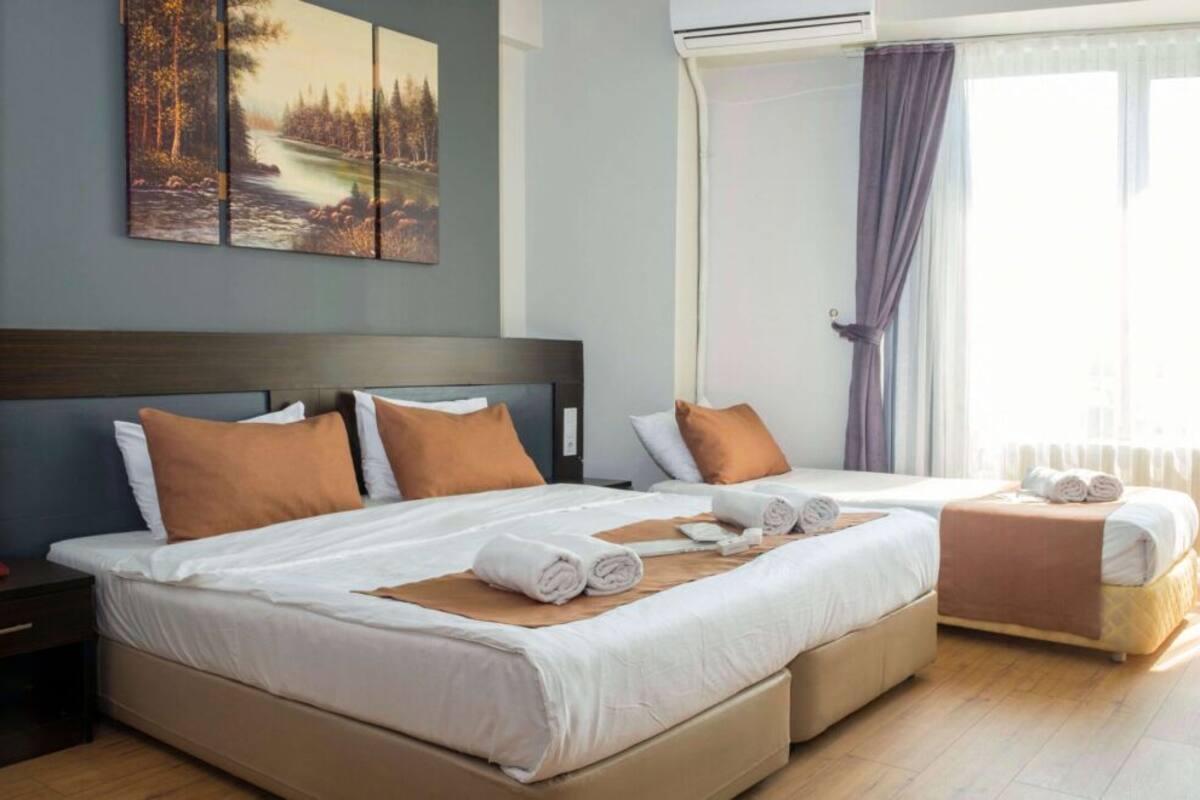 A hotel room with two beds, a large window with curtains, a painting on the wall, and a bedside table with a remote control.