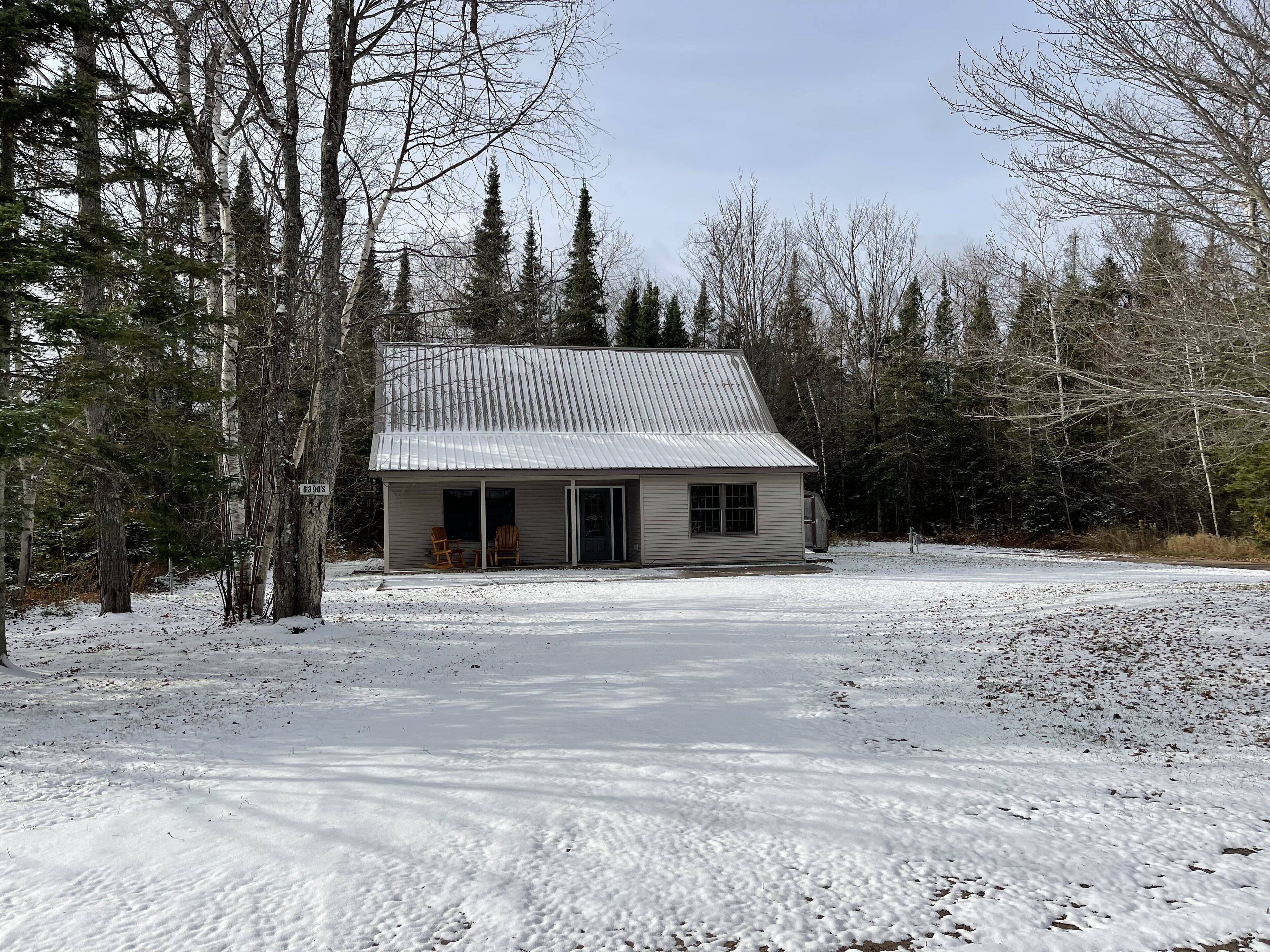 A small, single-story cabin with a metal roof, surrounded by a snowy landscape and dense forest.
