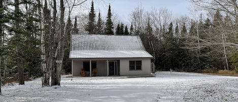 A small, single-story cabin with a metal roof, surrounded by a snowy landscape and dense forest.