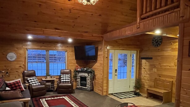 Ein gemütliches Blockhaus-Interieur mit Kamin, Ledersesseln, einem Holztisch und einem großen Fenster mit Jalousien.