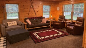 Ein gemütliches Blockhaus-Interieur mit einer braunen Ledersofa, einem passenden Sessel, einem Holztisch und einem rot-schwarzen Karotten Teppich.