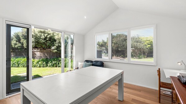 A room with a large table, a chair, a desk with a lamp, and a view of a garden through the windows.