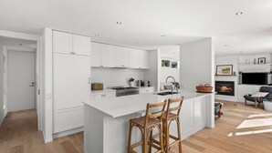 Uma cozinha moderna com armários brancos, uma ilha central com banquetas, fogão embutido e uma área de jantar com lareira.