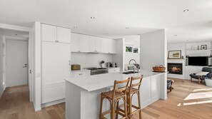 Et moderne køkken med hvide skabe, en central ø med barstole, et indbygget komfur og et spiseområde med pejs.