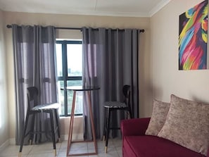 Une pièce avec un canapé rouge, deux fauteuils noirs, une petite table ronde et une fenêtre avec des rideaux gris.