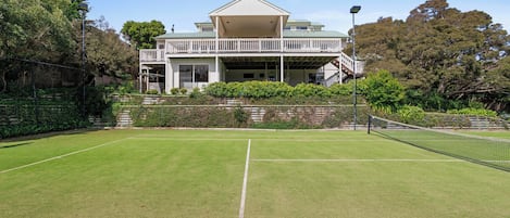 Una cancha de tenis con red, rodeada por una cerca, y un edificio con balcón al fondo.