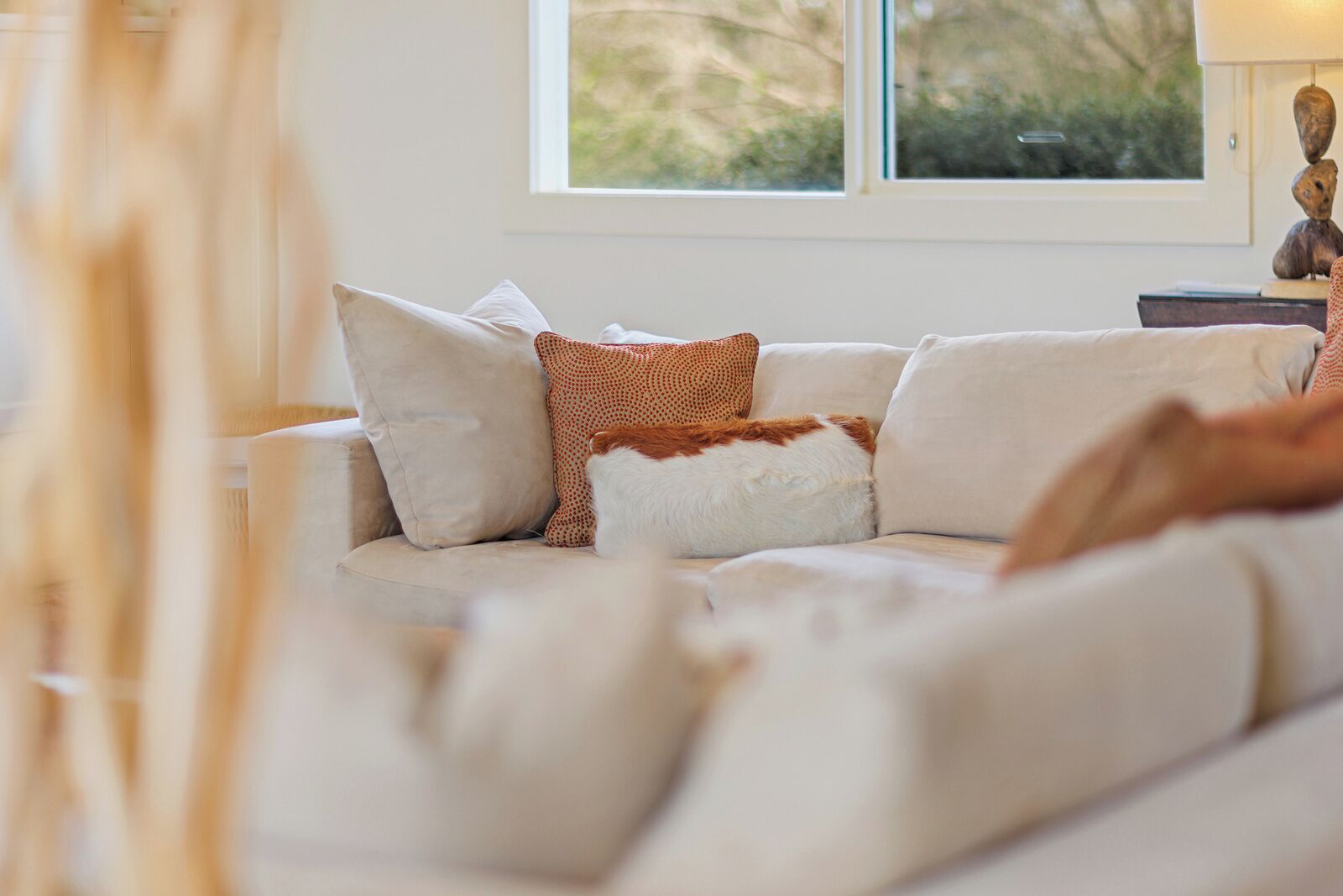 A modern living room with a beige sofa, patterned cushions, and a view of greenery through a window.