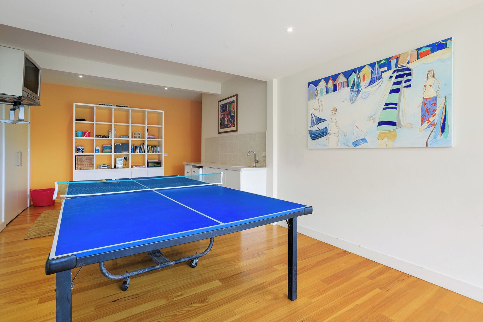 A room with a ping pong table, wooden flooring, a bookshelf, a television, and a painting on the wall.