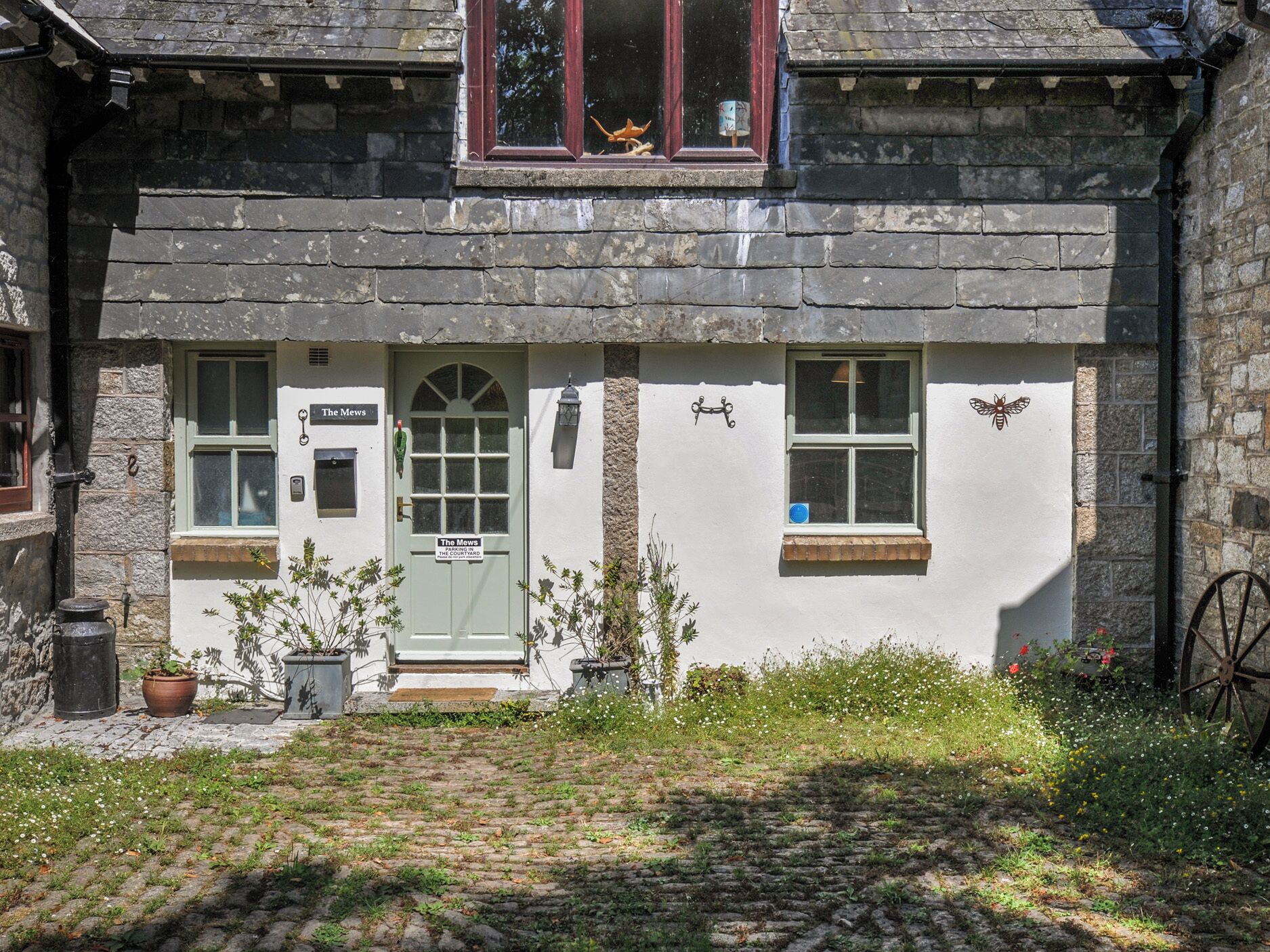 A traditional stone building with a green door, a sign reading 'The Mews,' and a small garden area with potted plants.