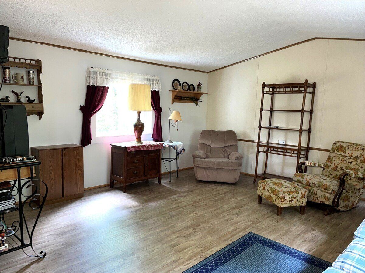 Una sala de estar con piso de madera, un televisor sobre un mueble bajo, un escritorio con lámpara, dos sillones, una escalera y un estante con objetos decorativos.
