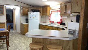 A compact kitchen with wooden cabinets, a white refrigerator, a coffee maker, and a bar area with stools. - Tree Top Retreat on the Peaceful Flambeau River! (Park Falls)