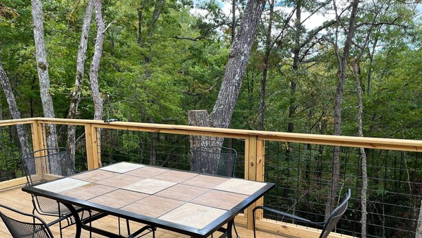 A wooden deck with a metal table and chairs, surrounded by a wooden railing and a view of a forest.