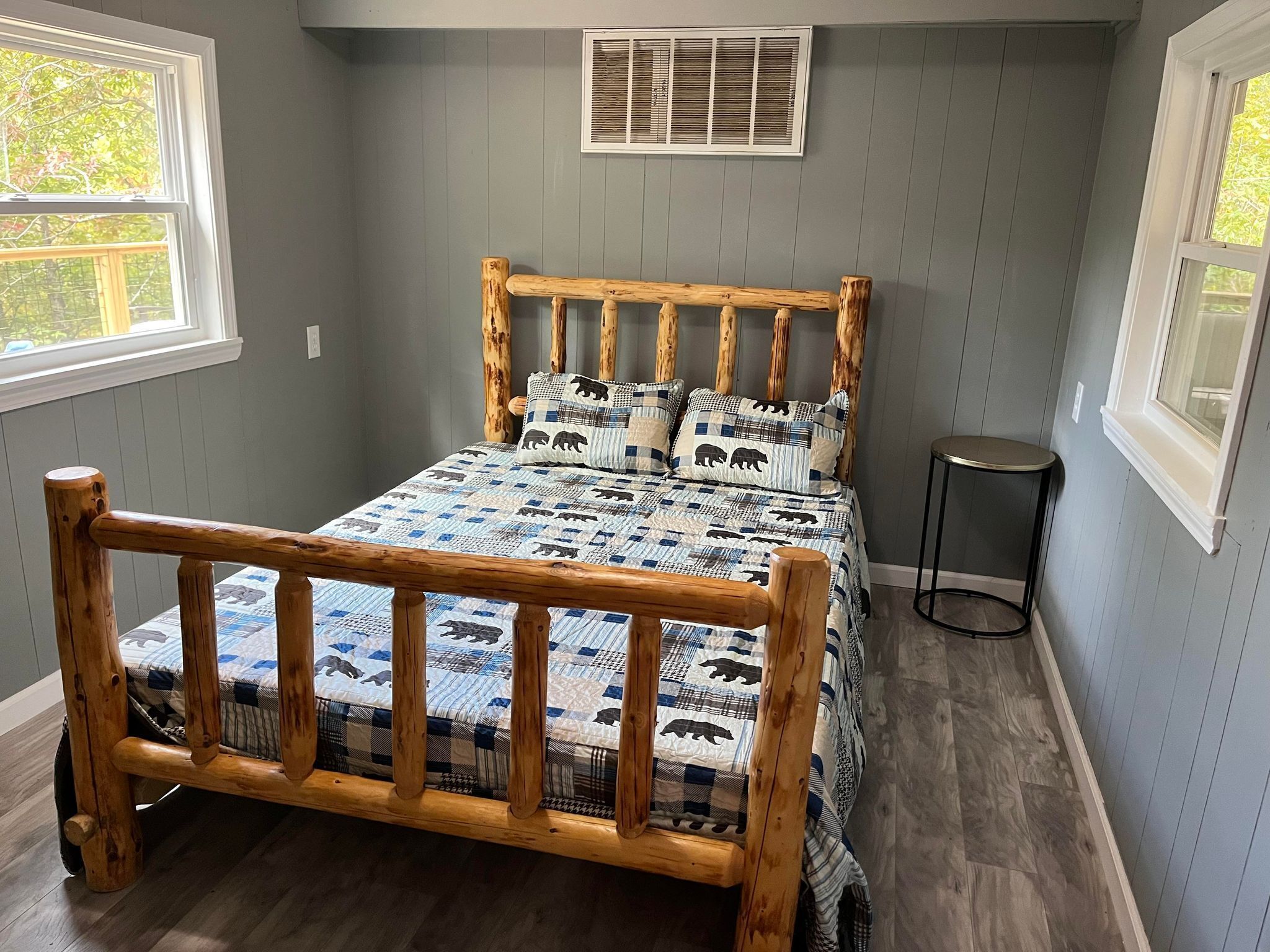A wooden bed with a checkered blanket, a small bedside table, and a window with blinds.