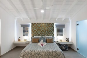 Une chambre à coucher avec une couvre-lit à motifs, deux tables de chevet avec des lampes, un ventilateur de plafond et un panneau décoratif mural.