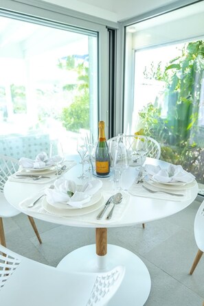Une table de salle à manger ronde, entourée de chaises blanches, mise pour un repas avec une bouteille de champagne, des verres et des couverts.