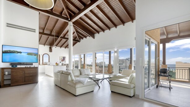 Amplia sala de estar con televisor grande, sofá y mesa de comedor. Hay un balcón con vista a la ciudad.