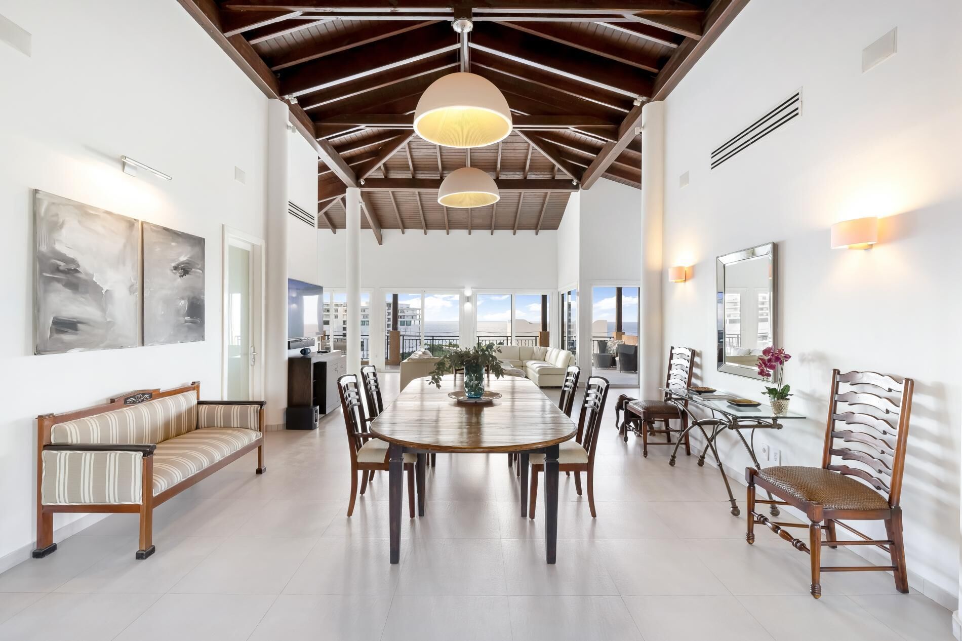 A spacious dining area with a high ceiling and wooden beams, featuring a large round table surrounded by chairs, a sofa, and a dining table with chairs.