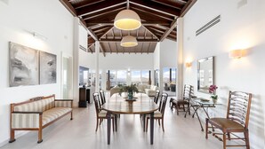 Un comedor amplio con techo alto y vigas de madera, que incluye una mesa redonda grande rodeada de sillas, un sofá y una mesa con sillas.