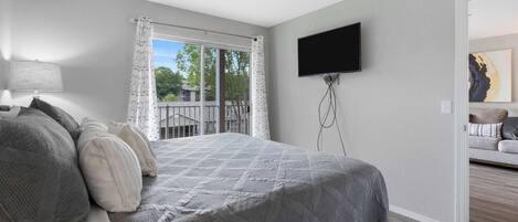 Une chambre avec un grand lit, un téléviseur fixé au mur et un balcon agrémenté de rideaux.
