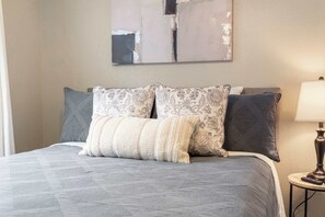 A bed with a quilted grey bedspread, two patterned pillows, a remote control on a bedside table, and a wall-mounted abstract painting. - The Branson Condo at Fall Creek - Overlooking the Rolling Hills (Branson)