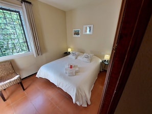 A neatly made bed with white linens, two pillows, and a set of towels on the floor. There is a window with a view of greenery outside, a chair, and two bedside tables with lamps. - Magnificent wooded villa, ideally located close to the beach. (L'Escala)