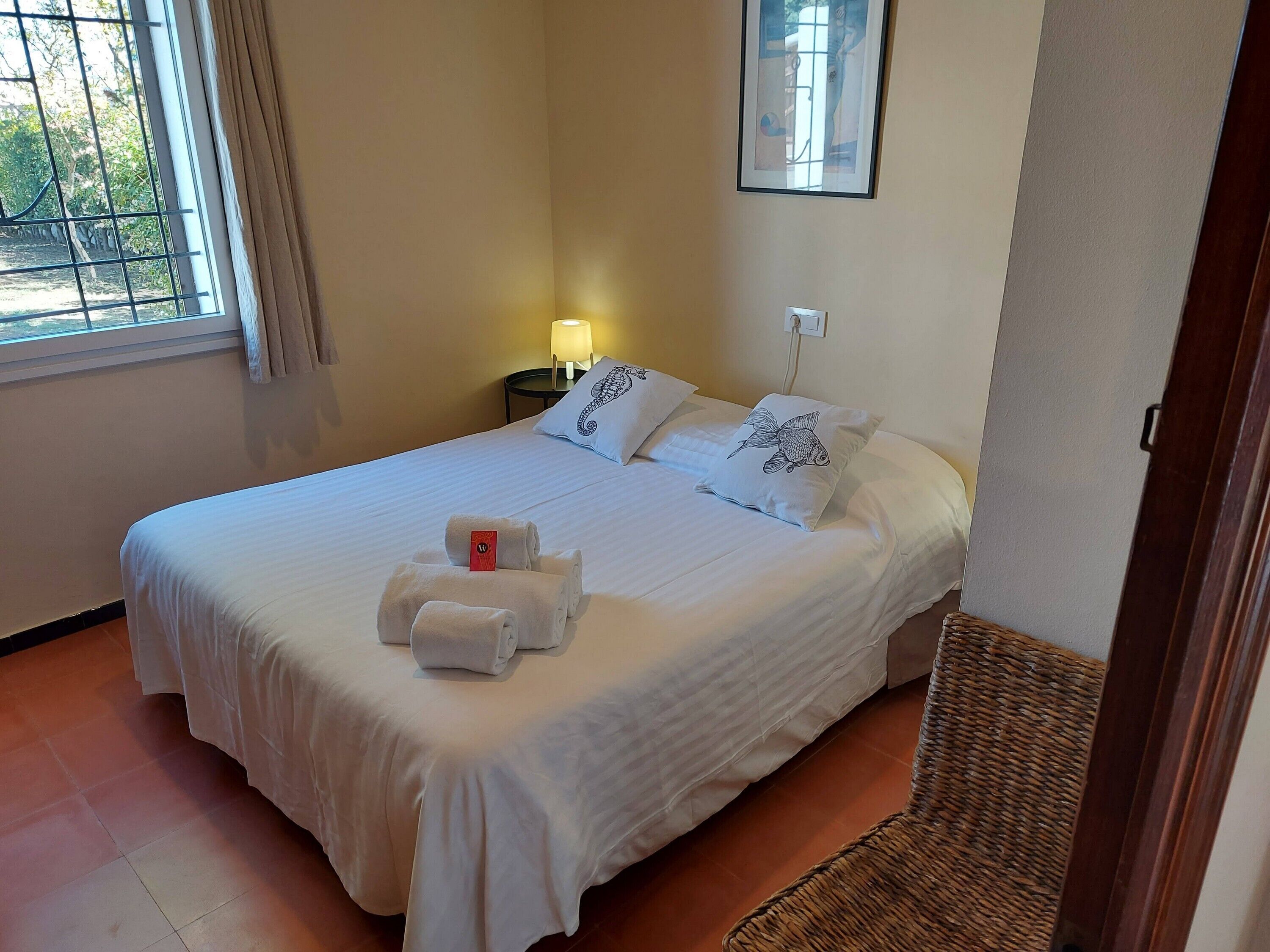 Een net opgemaakt bed met witte lakens en een kussen met een vispatroon, een klein nachtkastje met een lamp, en een raam met gordijnen.