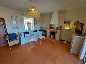 A living room with a stone fireplace, a sofa with cushions, a wicker chair, a small table, and a hanging light fixture. - Magnificent wooded villa, ideally located close to the beach. (L'Escala)