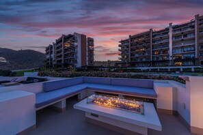 Et moderne udendørs loungeområde med bålsted, siddepladser og udsigt til høje bygninger.