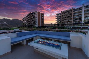 Een dak terras met een glazen vuurplaats, blauwe zitplaatsen en uitzicht op naburige gebouwen tijdens zonsondergang.