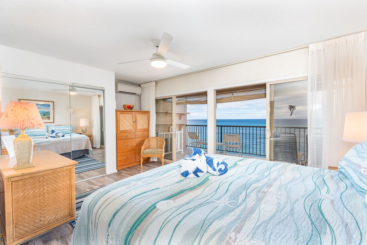 Hotelkamer met een bed, een dressoir, een stoel, een klein tafeltje, een plafondventilator en uitzicht op de oceaan door grote ramen.