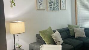 Ett modernt vardagsrum med en grå hörnsoffa, ett litet sidobord med en lampa och en hängande växt. På väggen hänger tre inramade botaniska tryck.