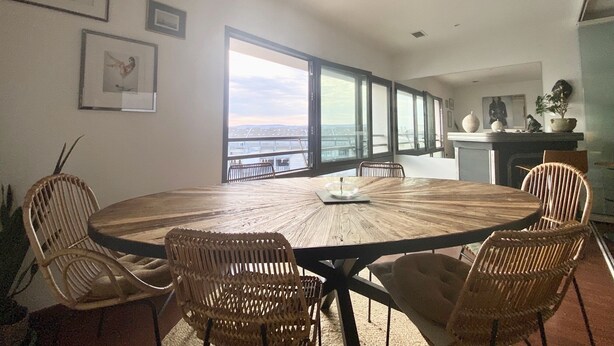 A round wooden table with wicker chairs, large windows with a view of a bridge, and framed pictures on the wall.