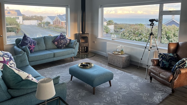 A living room with a sofa, ottoman, and a large window with a view of houses and greenery. - Watlands (Pwllheli)