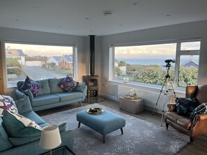 A living room with a sofa, ottoman, and a large window with a view of houses and greenery. - Watlands (Pwllheli)