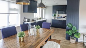 A modern kitchen with dark cabinets, pendant lighting, a wooden dining table, and potted plants. - Watlands (Pwllheli)