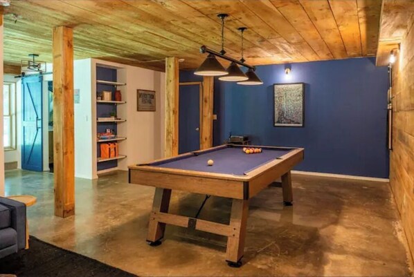 Une table de billard dans une pièce dont le plafond est orné de poutres en bois, avec des étagères remplies d’objets et un mur bleu.