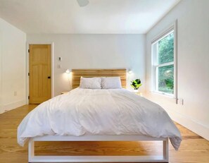 Une chambre à coucher avec une tête de lit en bois, du linge de lit blanc, une plante sur la table de chevet et une grande fenêtre donnant sur un paysage verdoyant.
